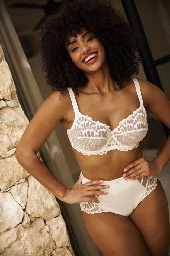 Model in a white lace underwired bra and high-waisted brief, standing by a stone wall.