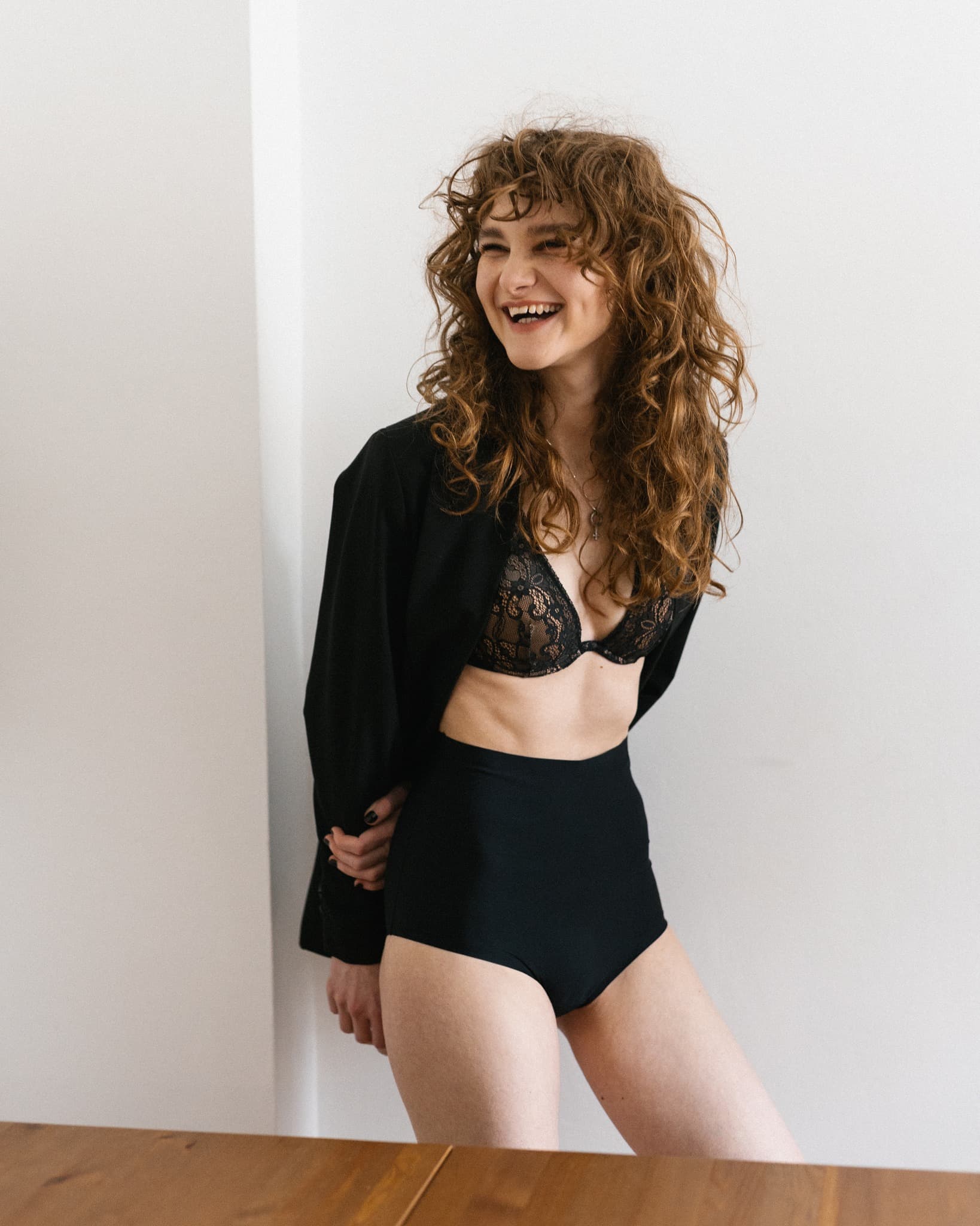 Model in a black lace bra and high-waisted brief under an open black jacket, laughing.