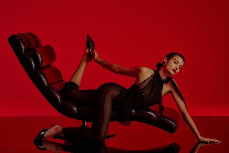 Model in sheer black mesh, reclining on a black leather lounge chair against a red background.