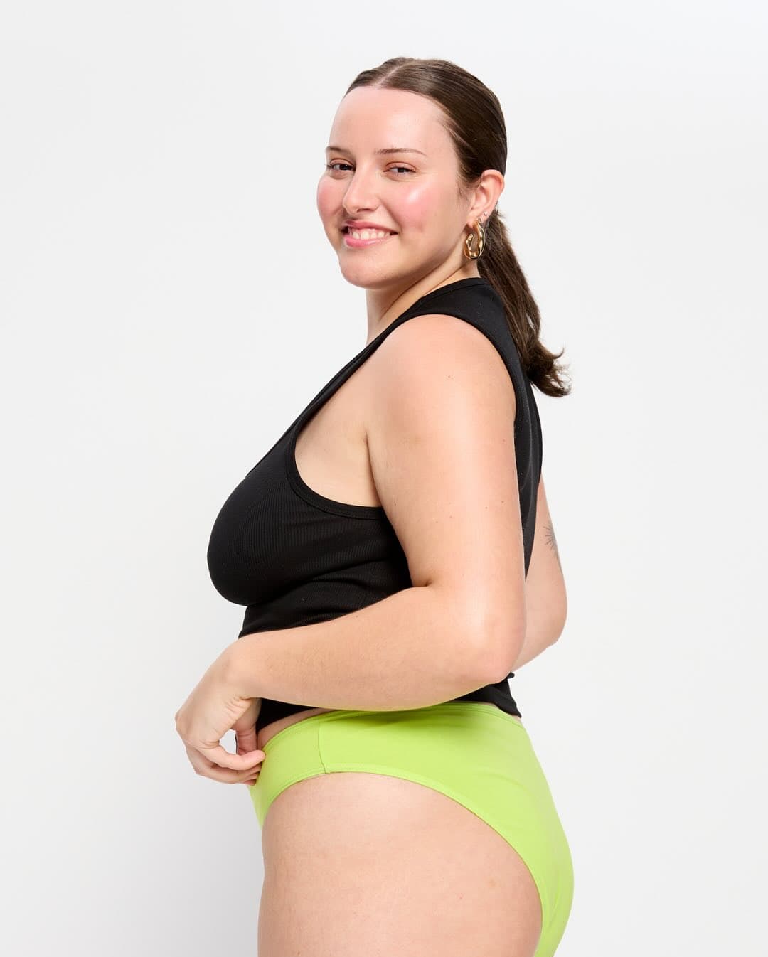 Model in a black tank top and lime green period brief, seen from the side against a white background.