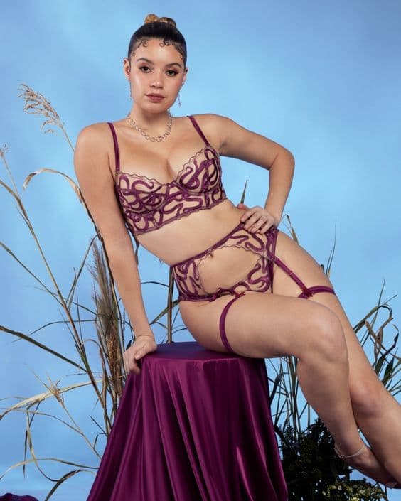Model in a burgundy bra and suspender set with sculpted appliqué, sitting on a draped stool against blue.