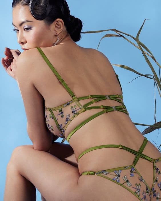 Model seen from the side in a green strappy bra and brief with embroidered mesh, against a blue sky.