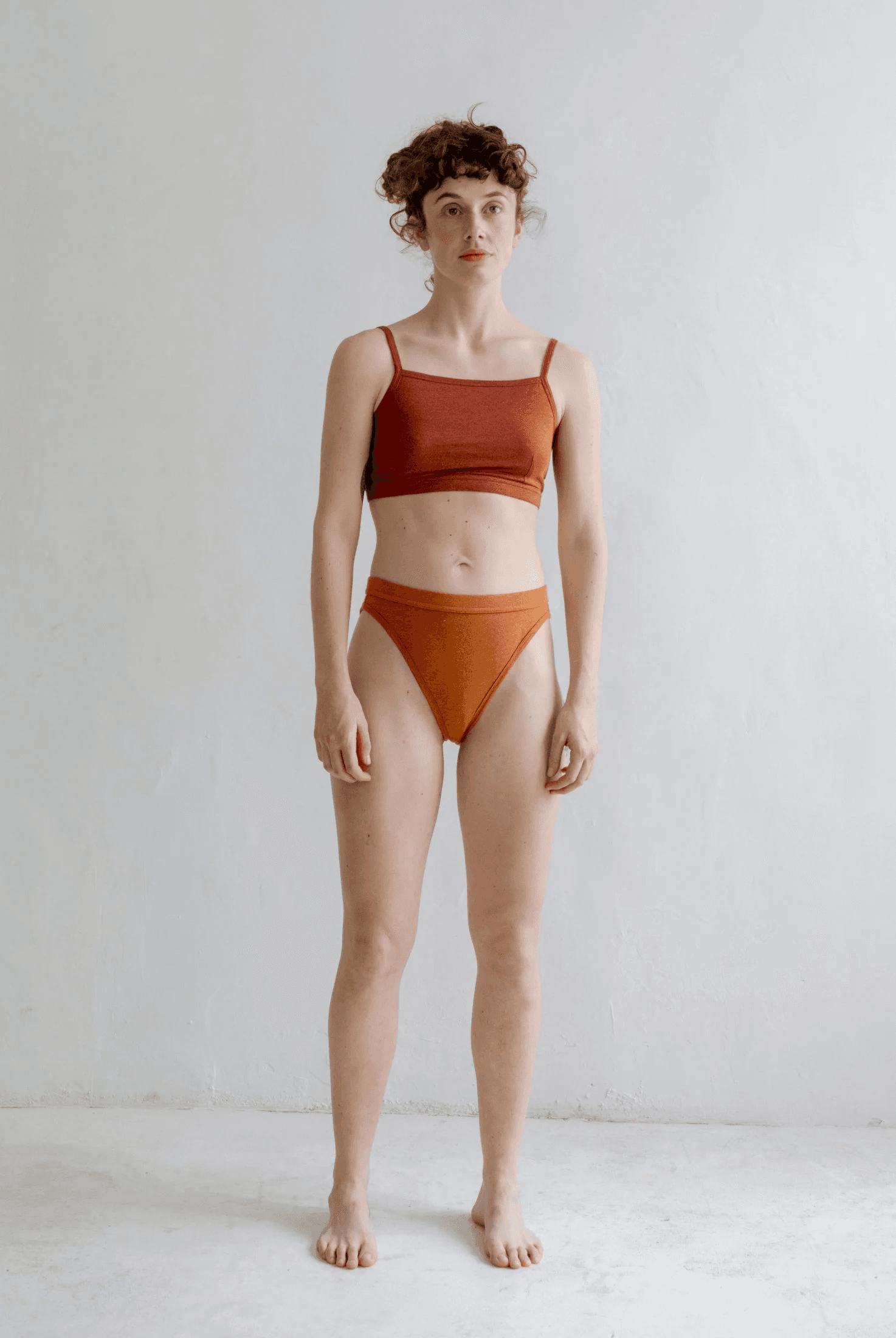 Model in a rust bandeau bralette and orange high-waisted brief, standing barefoot against a white wall.